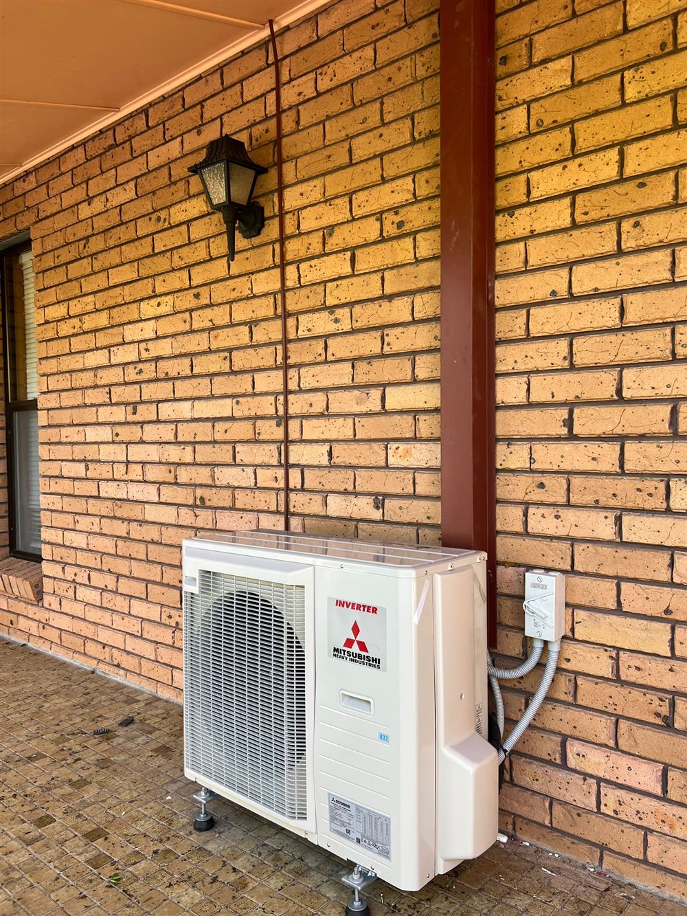 Outdoor air conditioning unit installed on adjustable feet beside a brick wall with a tidy isolator switch